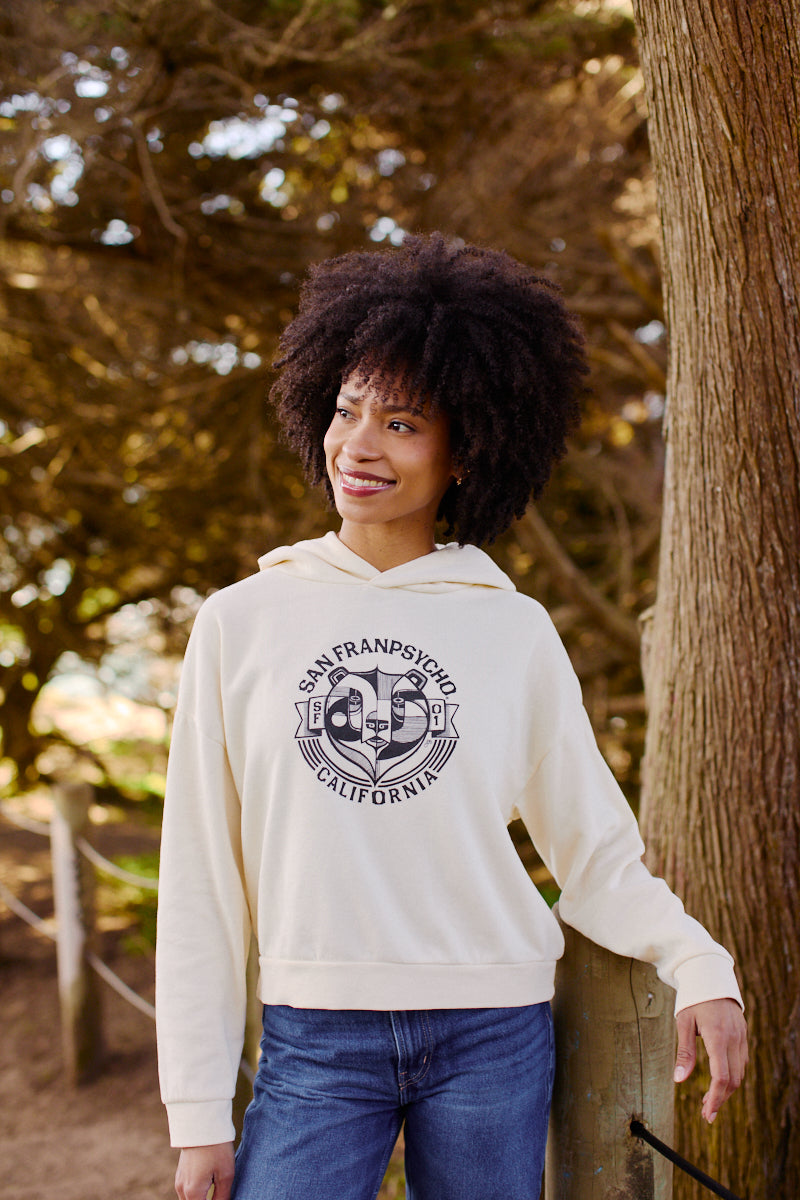 A woman in a Women's Bear Essentials Pullover and jeans smiles outdoors by a tree, sunlight streaming behind her.