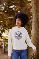 A woman in a Women's Bear Essentials Pullover and jeans smiles outdoors by a tree, sunlight streaming behind her.