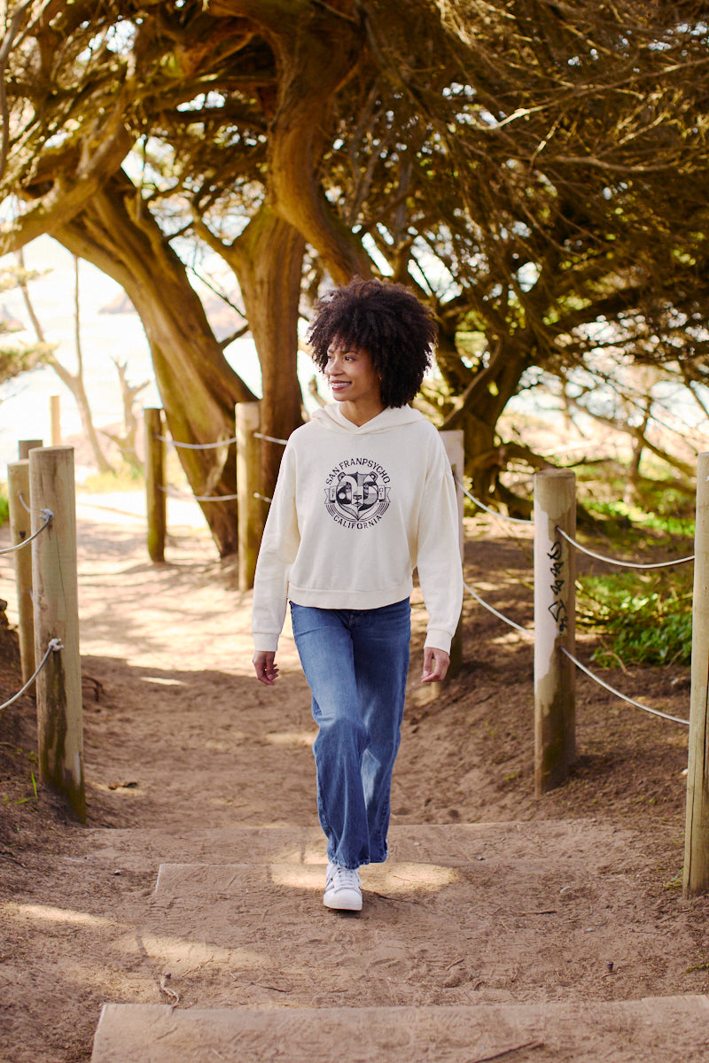 A person with curly hair walks along a sandy path lined with trees, wearing jeans and the ultra-soft Women's Bear Essentials Pullover.