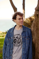 A young man in a denim jacket leans against a tree, smiling, with his Bear Essentials Tee in Heather Grey peeking out—perfect for relaxing by the greenery and water.