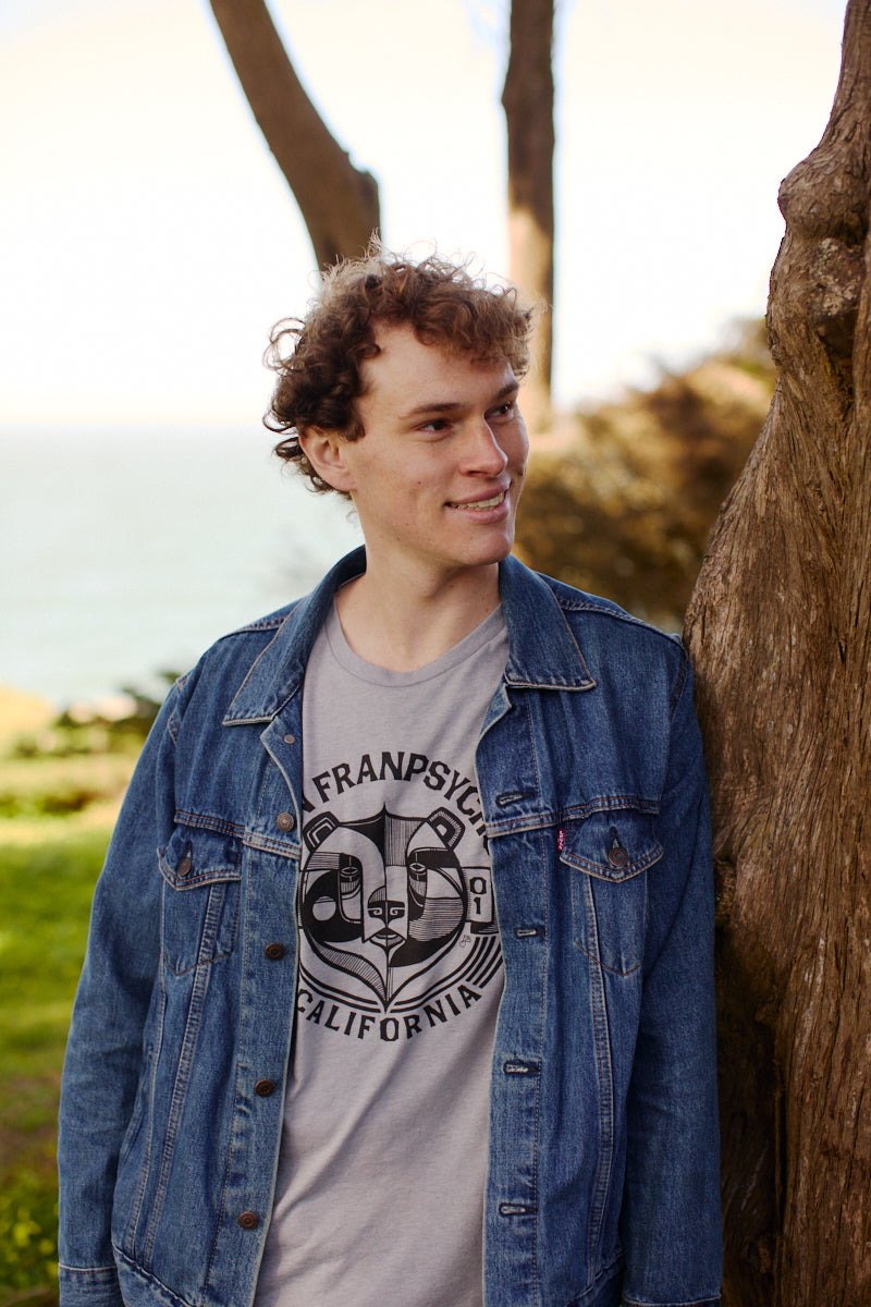 A young man in a denim jacket leans against a tree, smiling, with his Bear Essentials Tee in Heather Grey peeking out—perfect for relaxing by the greenery and water.
