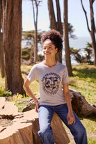 A woman smiles outdoors in a forest, wearing the ultra-soft Bear Essentials Tee - Heather Grey with a California bear graphic and jeans.