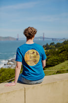 Wearing the Elemental Tee - Blue, a person sits on a ledge overlooking the Golden Gate Bridge and San Francisco Bay on a sunny day.