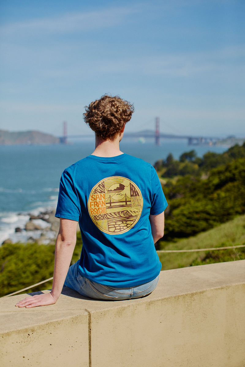 Wearing the Elemental Tee - Blue, a person sits on a ledge overlooking the Golden Gate Bridge and San Francisco Bay on a sunny day.