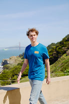 A young man wearing the Elemental Tee - Blue strolls outdoors with the Golden Gate Bridge and lush greenery in the background, embodying San Francisco’s relaxed vibe.