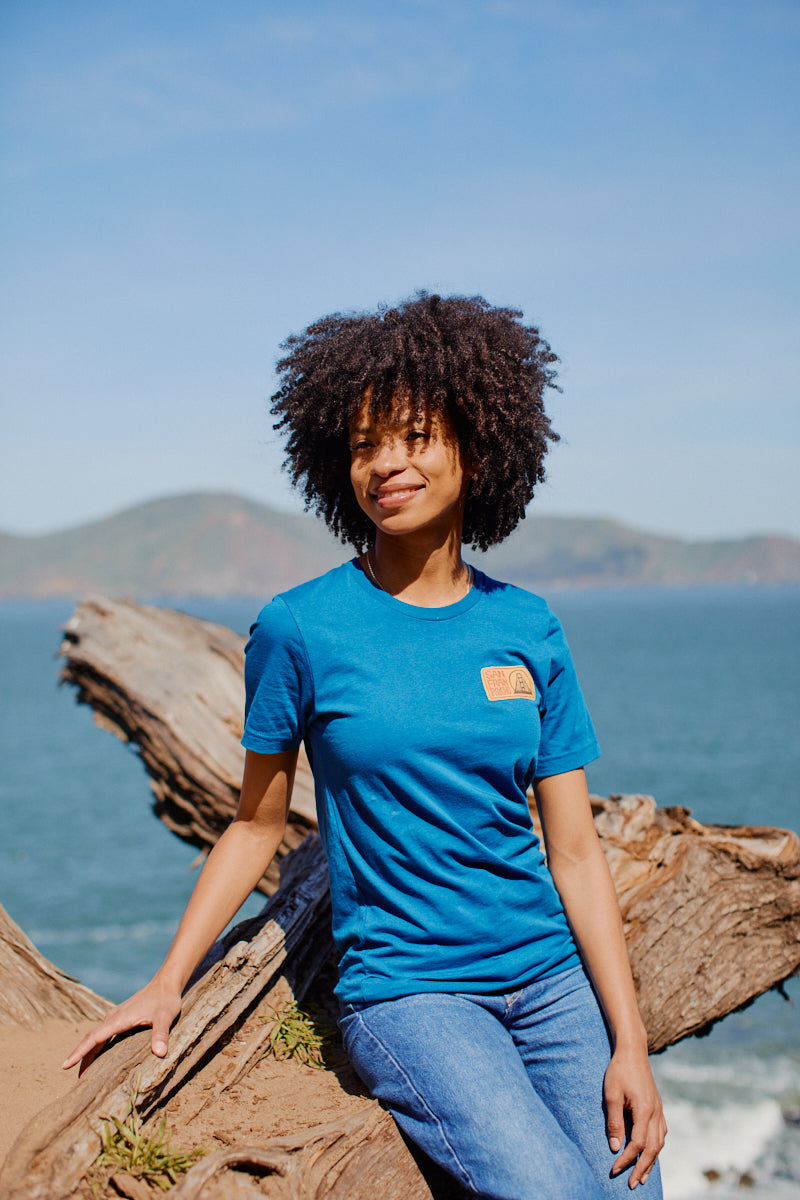 A woman in the Elemental Tee - Blue and jeans sits on driftwood by the sea, with San Francisco hills and a clear sky in the background.