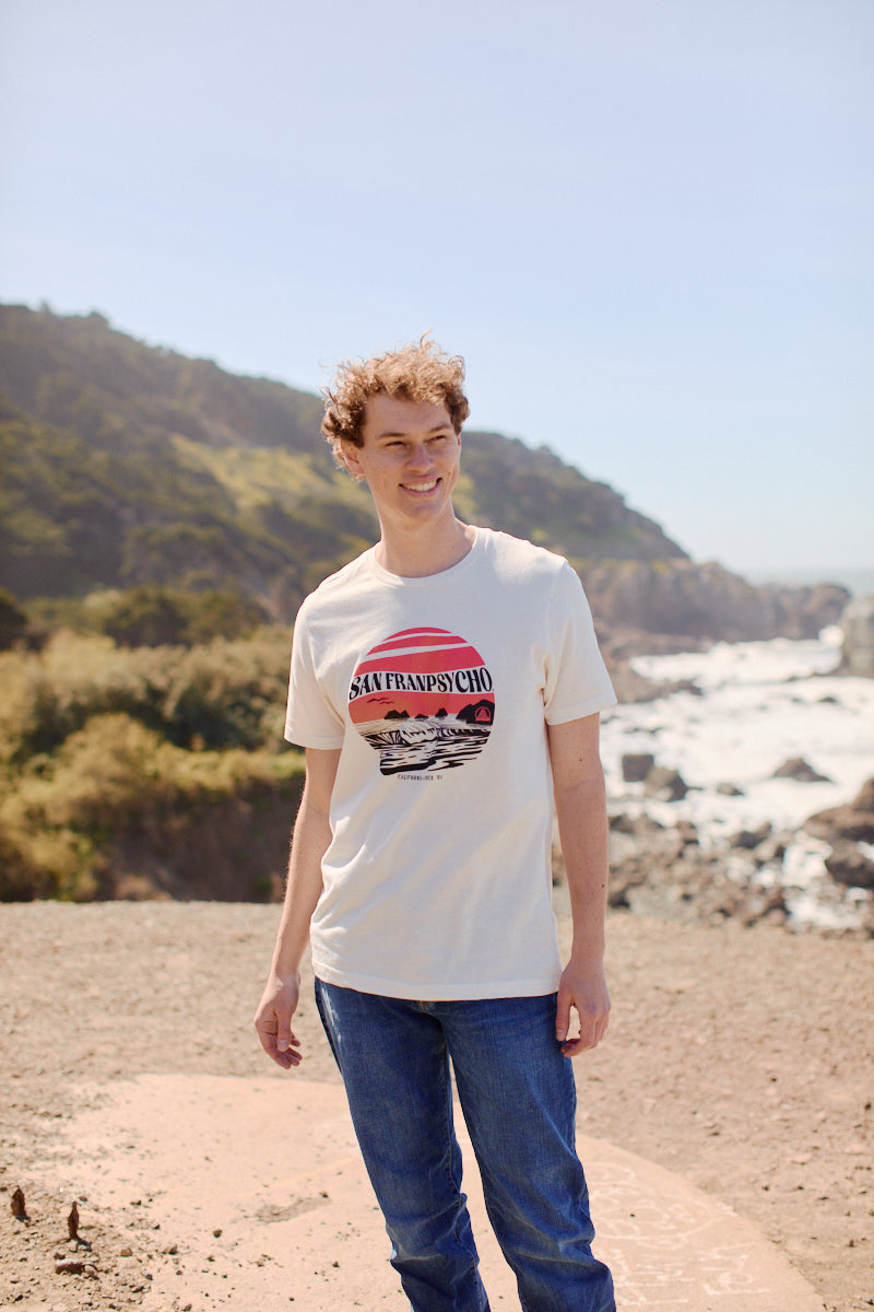 A smiling person in jeans and a Kelly's Cove Tee - Vintage White stands outdoors by the ocean and cliffs, enjoying the sunny coastal city vibes.