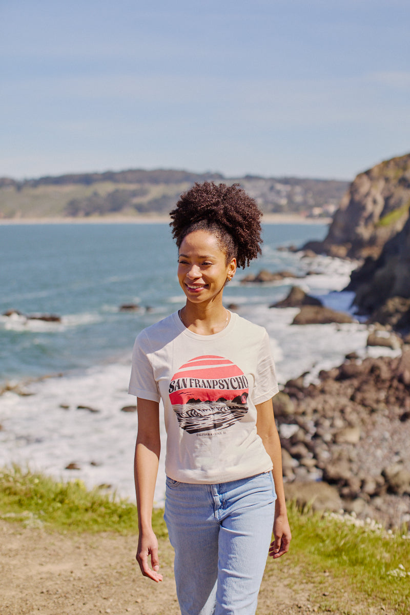 A woman in the Kelly's Cove Tee - Vintage White strolls along a rocky coastline, ocean and cliffs behind her—capturing relaxed coastal city style.