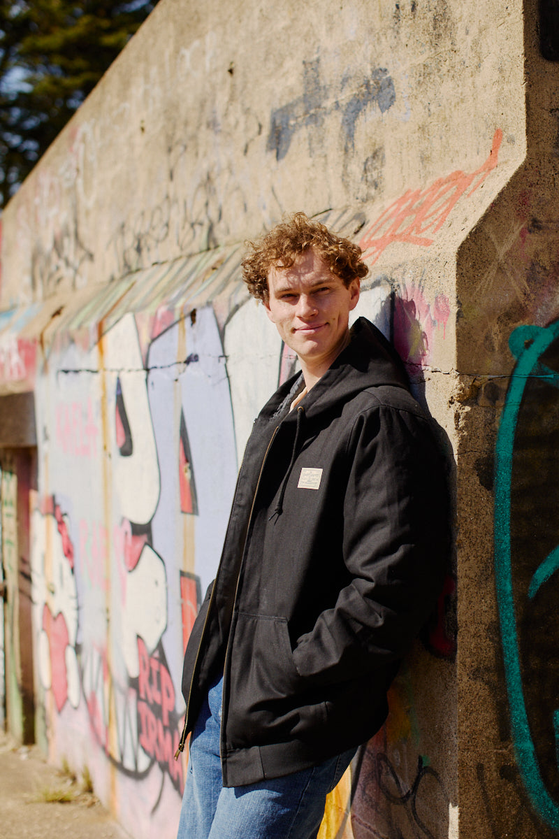 A young man smiles on a sunny day, leaning against a graffiti-covered wall while wearing the Bridge Co. Workwear Jacket in black cotton canvas.