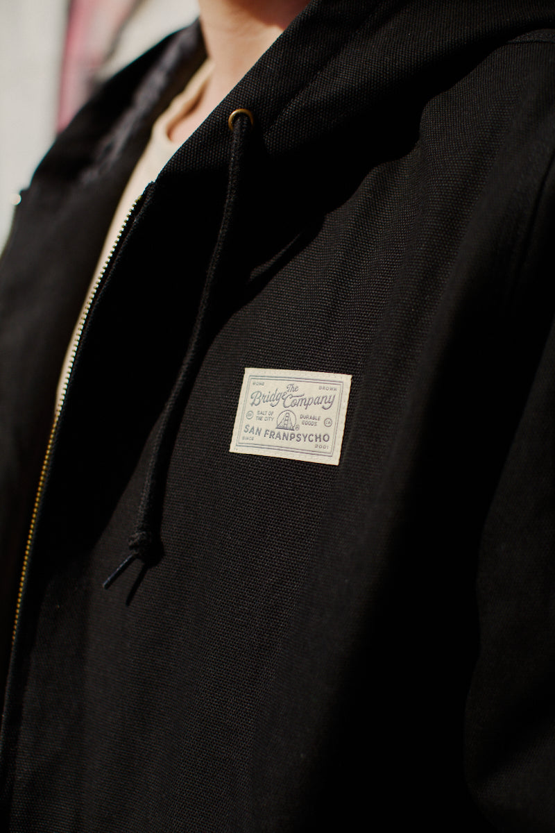 Close-up of the Bridge Co. Workwear Jacket in black cotton canvas, featuring a zip-up front and a white patch that reads The Daily Company San Francyscho.
