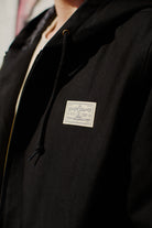 Close-up of the Bridge Co. Workwear Jacket in black cotton canvas, featuring a zip-up front and a white patch that reads The Daily Company San Francyscho.