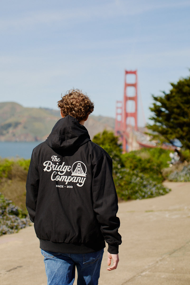 A person in a black hoodie and the Bridge Co. Workwear Jacket walks near the Golden Gate Bridge in daylight.