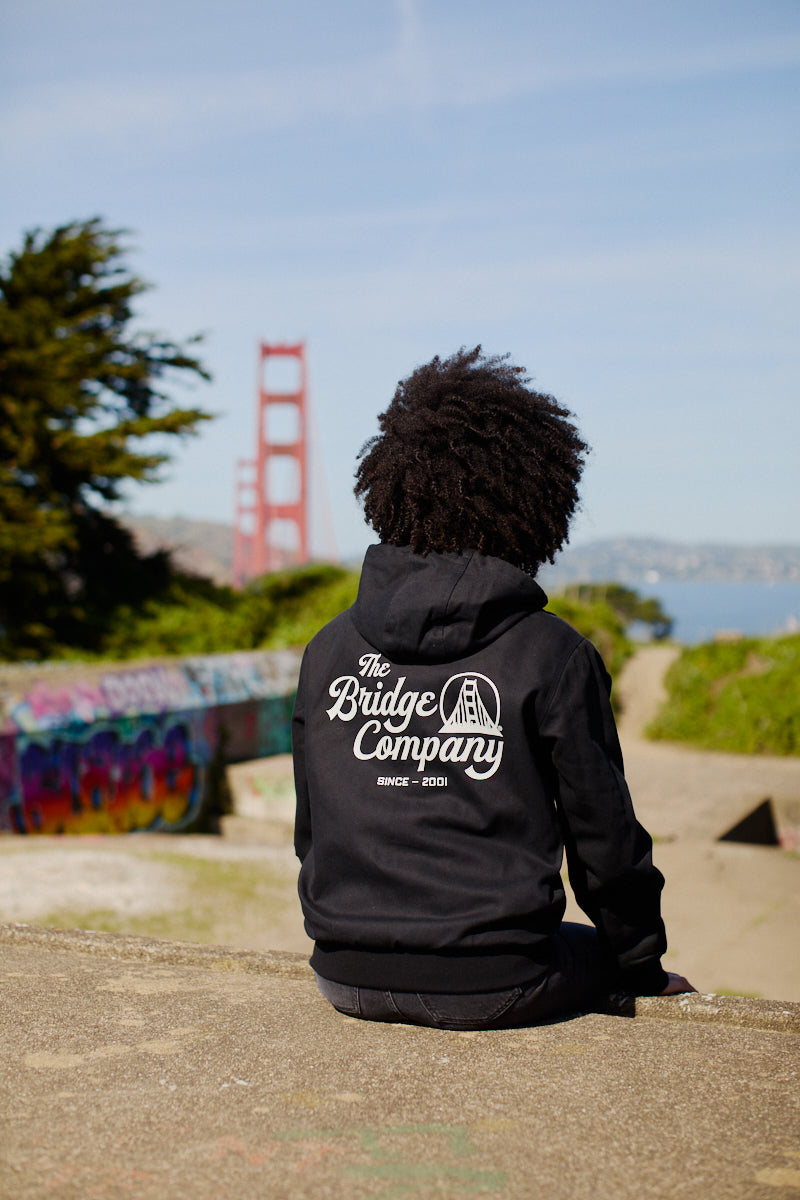 Wearing a Bridge Co. Workwear Jacket, a person sits outdoors on a sunny day, facing the Golden Gate Bridge.