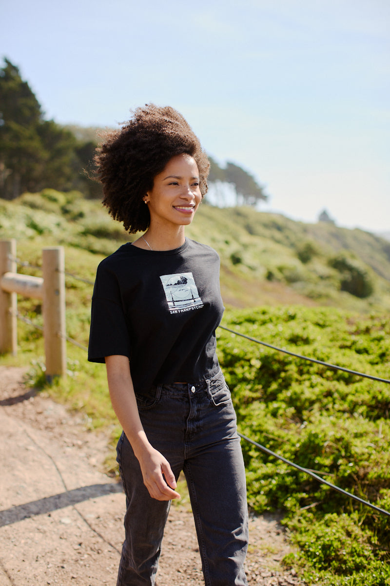 A smiling woman strolls along a scenic, sunlit path in greenery and distant trees, wearing the Women's Elemental Cropped Tee in black with its bold graphic design.