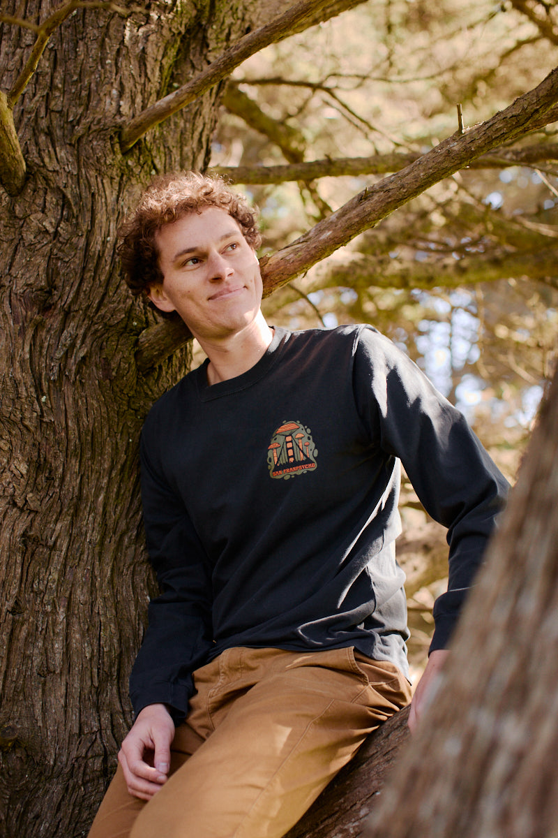 A person with curly hair sits on a tree branch, wearing the Mind Bloom Long Sleeve inspired by Erik Abel artwork and tan pants.
