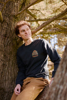 A person with curly hair sits on a tree branch, wearing the Mind Bloom Long Sleeve inspired by Erik Abel artwork and tan pants.