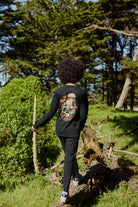 Wearing the Mind Bloom Long Sleeve, a person in black walks on logs through a sunlit, green forest with tall trees in the background.