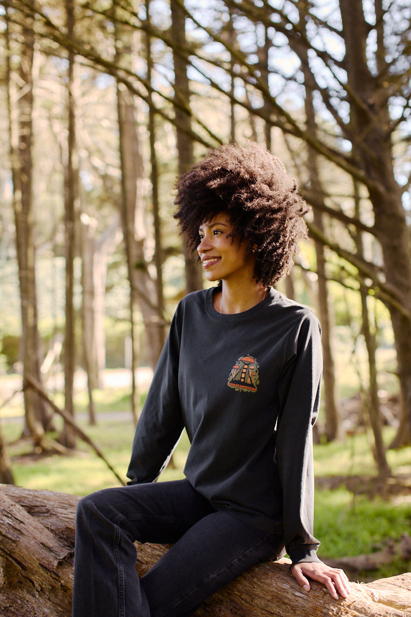 A woman with curly hair smiles while sitting on a fallen tree in the forest, wearing the Mind Bloom Long Sleeve, a graphic tee inspired by Erik Abel's artwork.