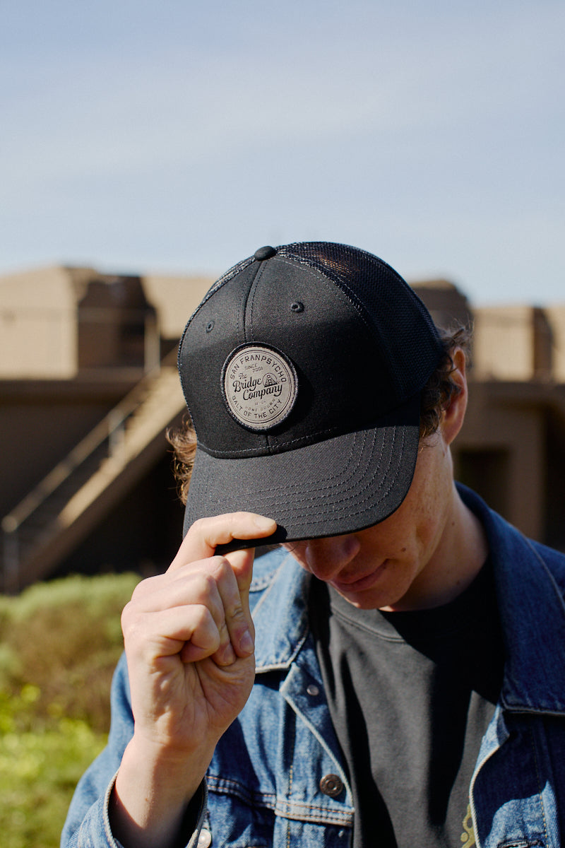 Outdoors on a sunny day, a person in a denim jacket tips the Salt of The City Trucker Hat—a black hat featuring a round patch and adjustable back.