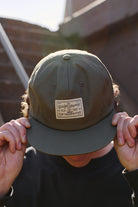 Person in a dark shirt tipping the Bridge Co. Patched Hat, featuring a green cap with a "The Bridge Company San Franpsycho" patch, adjustable back for comfort, and one size fits most design.