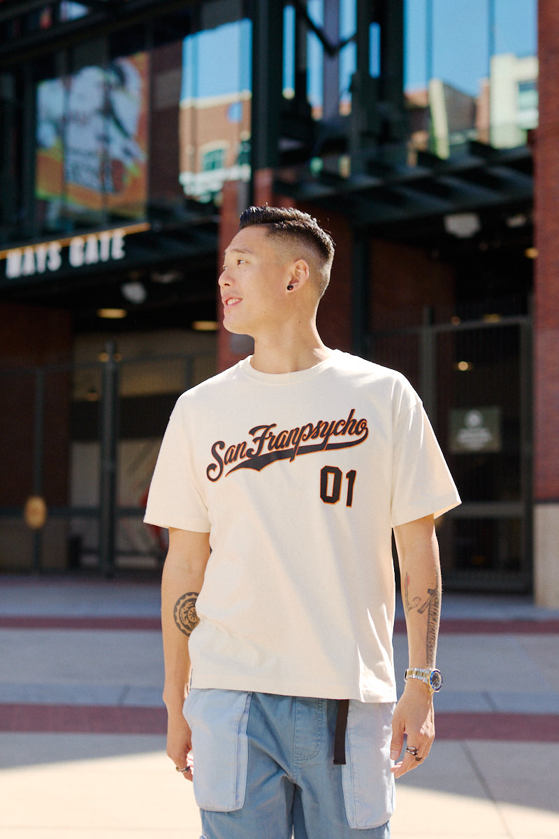 A man in a Psychos Script Tee - Natural stands outside a stadium gate, looking to the side.