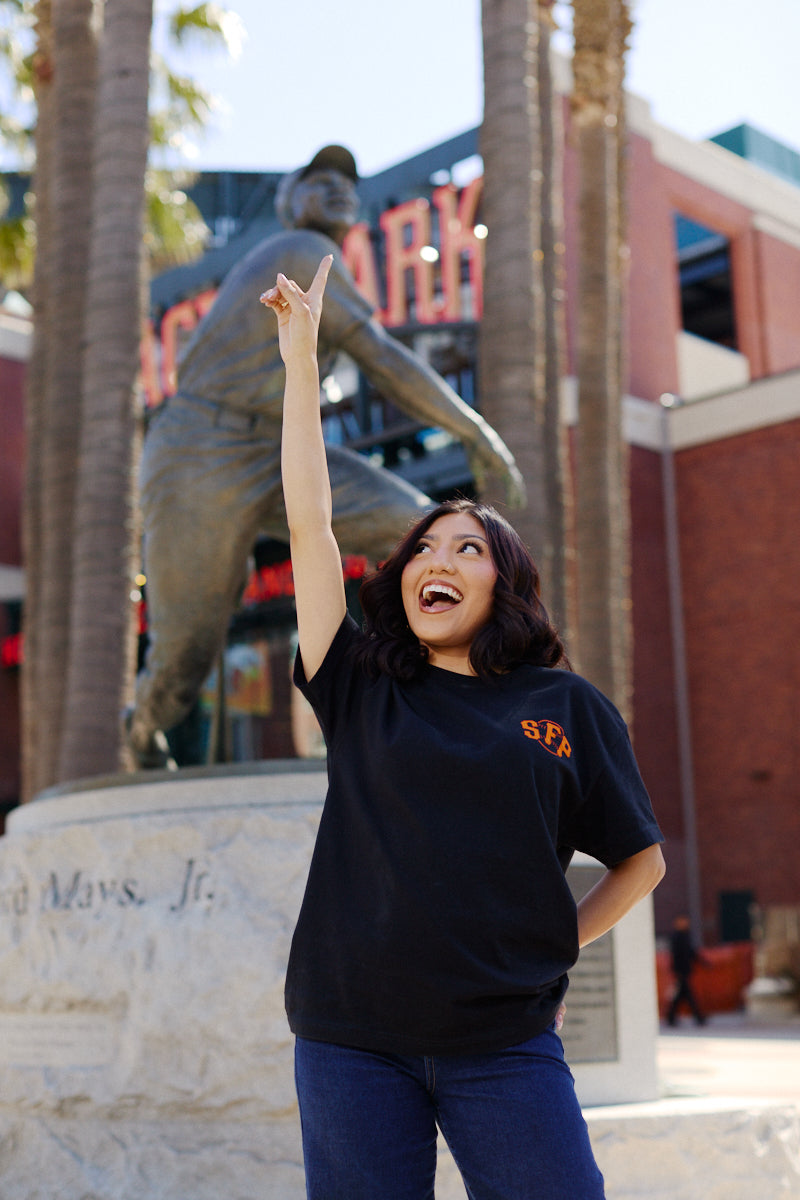 A woman smiles and points up in front of a giant baseball player statue outside the stadium, proudly wearing the Black & Orange Psychos Tee.