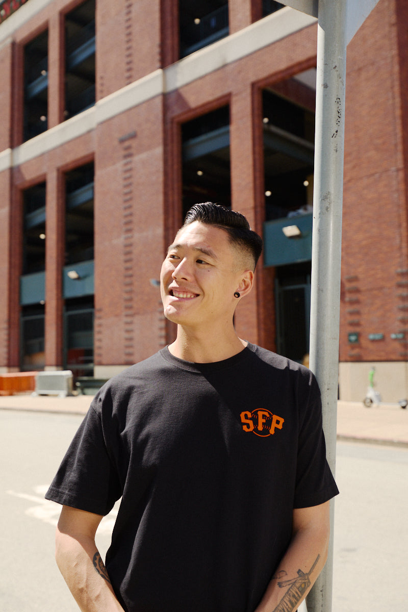 A person smiles while standing outside near a brick building and a street sign, wearing the Black & Orange Psychos Tee.