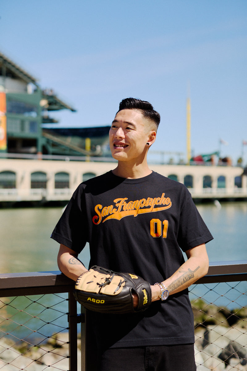 A man wearing the Psychos Script Tee - Black, featuring hand-printed orange graphics, holds a baseball glove while standing by a waterfront stadium.