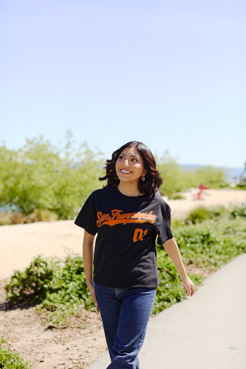 A woman smiles while walking outdoors on a sunny day, wearing the Psychos Script Tee - Black with a hand-printed San Francisco 01 design in black and orange, paired stylishly with jeans.