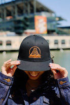 Smiling person outdoors, holding the brim of a black & orange Embroidered Logo Snapback with an adjustable back and bridge logo.
