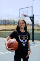 A smiling young woman wears a Valhalla Tee while holding a basketball on an outdoor court, with a hoop and net in the background—show your Valkyrie spirit this season with the Valhalla Tee.