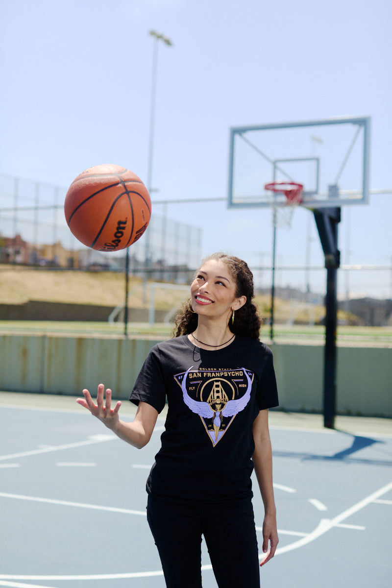 A woman smiles and spins a basketball on her finger at an outdoor court, wearing the Valhalla Tee—crafted from soft Airlume cotton and ideal for showing your Valkyries spirit this season.
