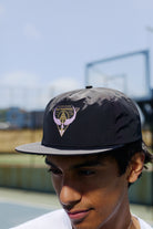 A young man wears a black Valhalla Hat with an adjustable back outdoors near a tennis or basketball court.