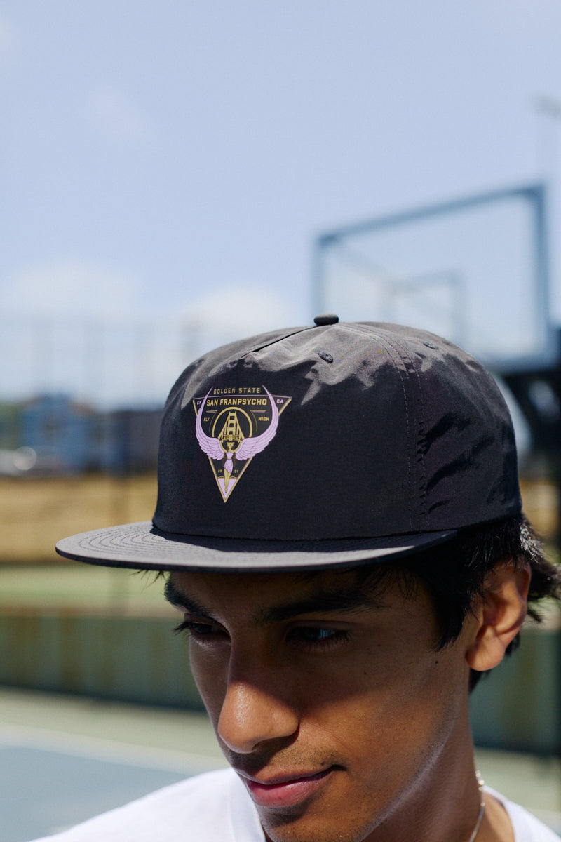 A young man wears a black Valhalla Hat with an adjustable back outdoors near a tennis or basketball court.