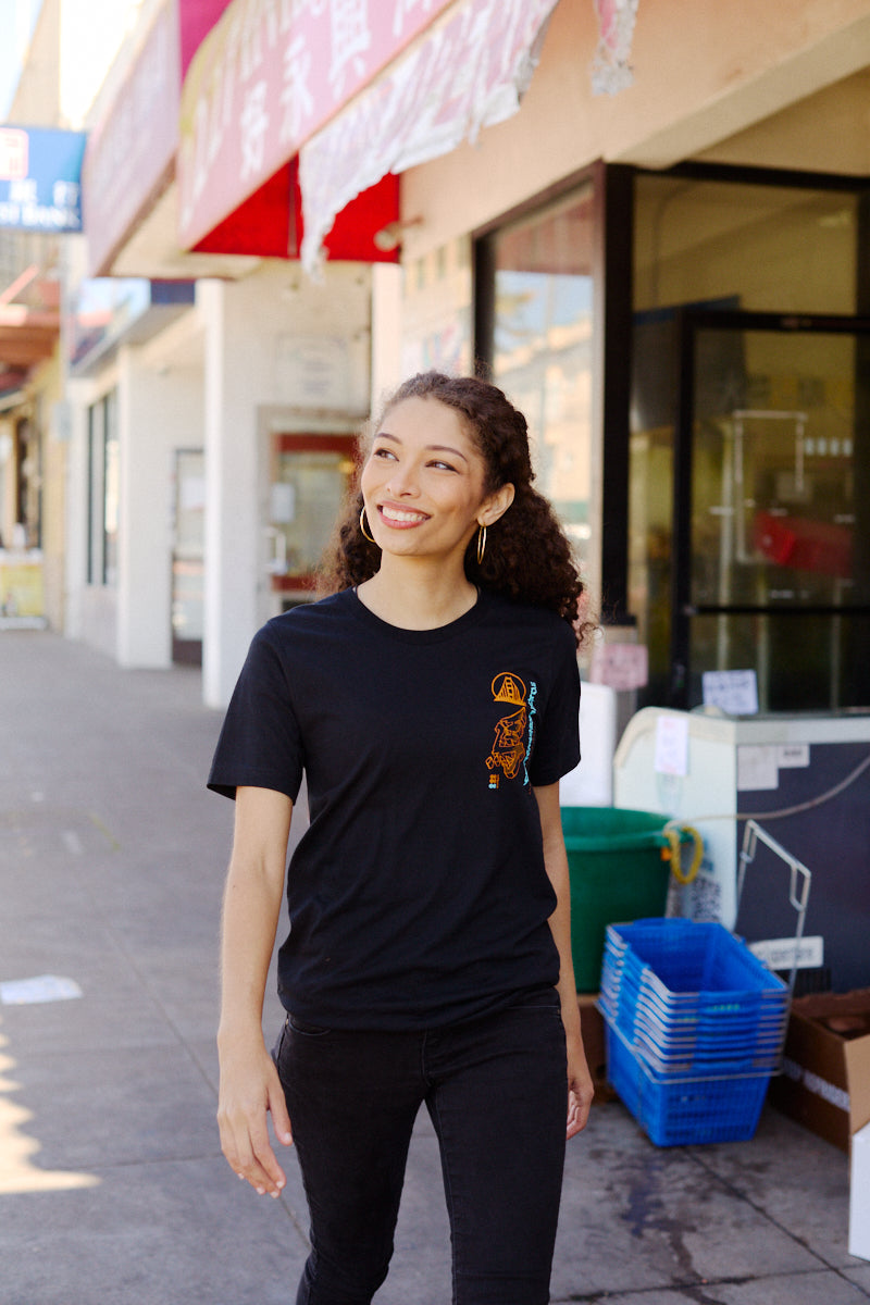 A woman wearing the Year of the Horse Tee walks on a sunny sidewalk lined with shops displaying Chinese signs, celebrating Lunar New Year.