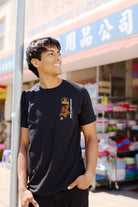 A smiling young man wearing the Year of the Horse Tee stands outside a store with colorful items and Chinese signage, celebrating Lunar New Year.