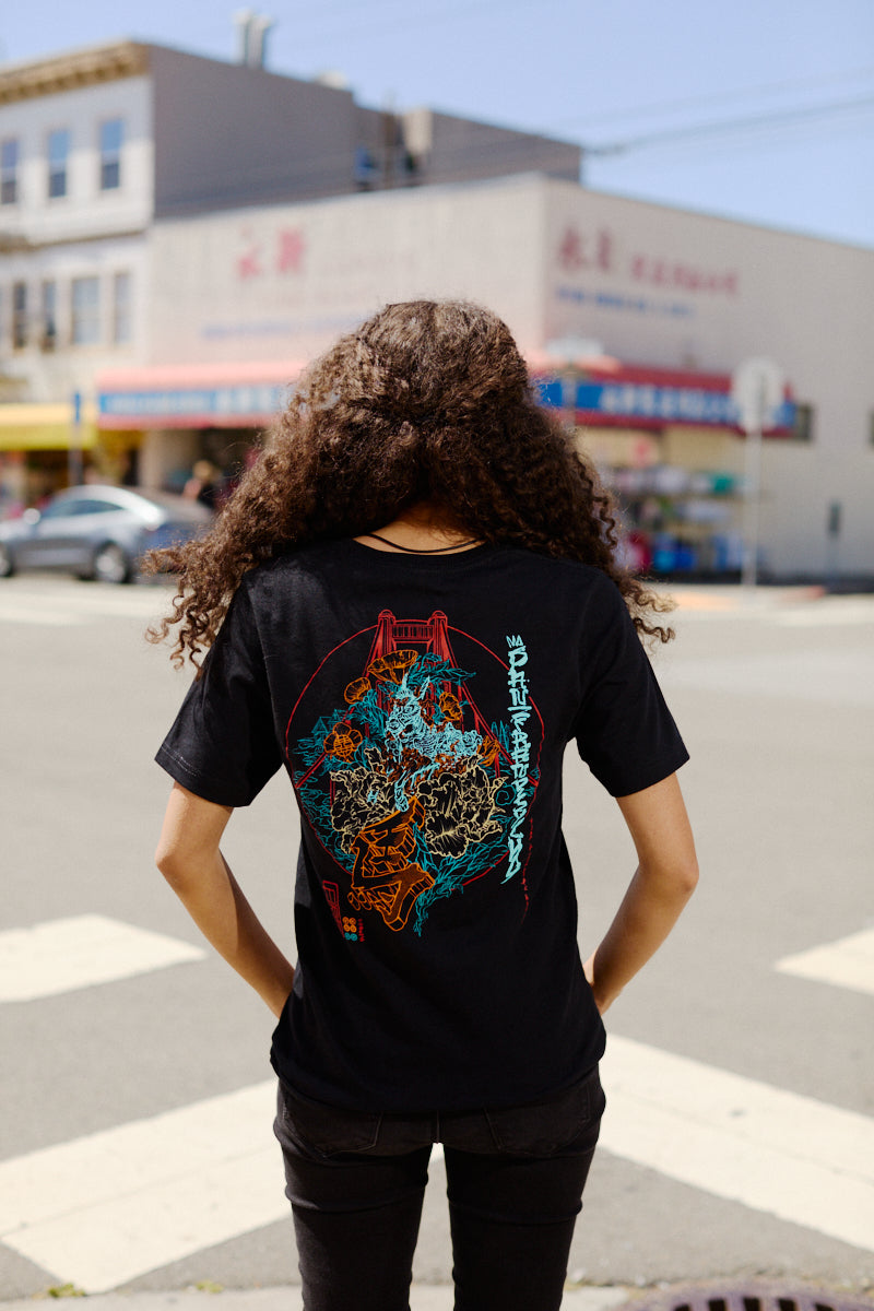 A person with curly hair wearing a Year of the Horse Tee stands at a crosswalk in a city neighborhood.