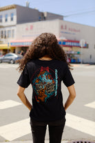 A person with curly hair wearing a Year of the Horse Tee stands at a crosswalk in a city neighborhood.