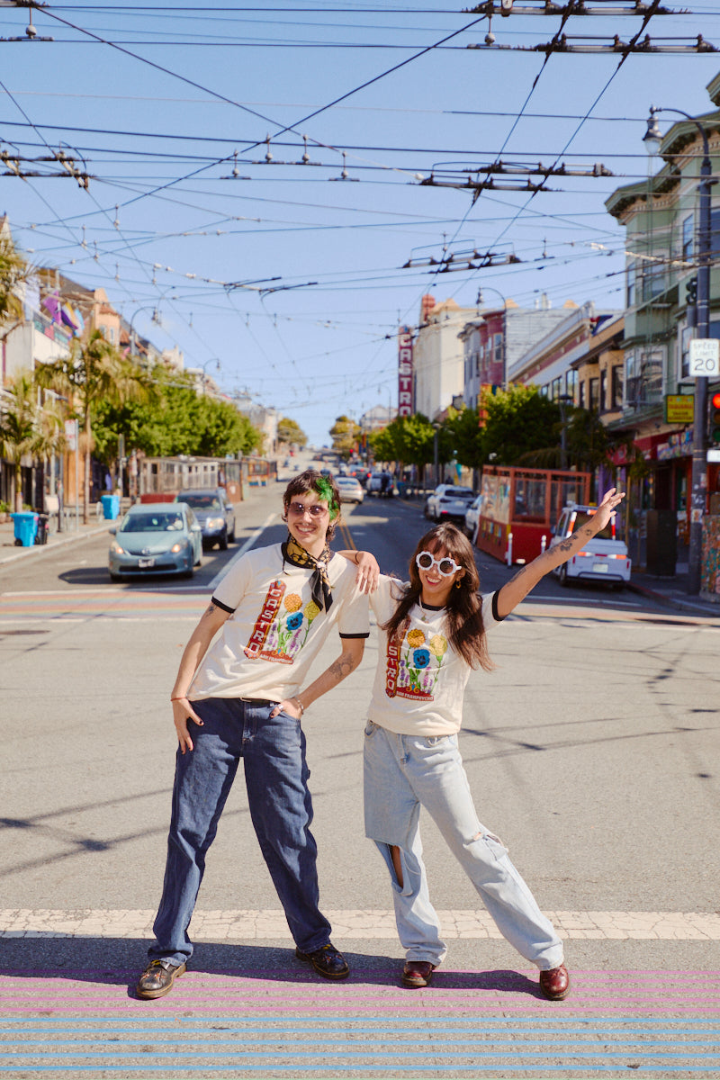 Two people in Castro Pride Tees and sunglasses pose playfully on a sunny city street near Castro Theatre, their vibrant shirts standing out amid the lively city backdrop.