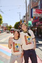 Two smiling people wearing Castro Pride Tees stand on a sunny, rainbow-striped street in San Francisco, showcasing the shirt's bold Pride design.