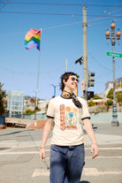 A person wearing the Castro Pride Tee smiles on a sunny street, rainbow flag and iconic Castro Theatre sign in the background, celebrating with queer flowers.