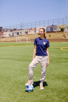 Woman on a soccer field in San Francisco, one foot on a blue and white soccer ball, goalposts behind her—sporting the WC x SFP Tee and ready to represent Team USA at the World Cup.