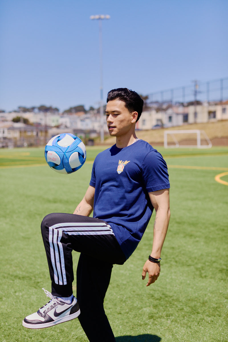 Wearing the WC x SFP Tee, a man balances a blue and white soccer ball on his knee in sunny San Francisco, channeling Team USA’s energy as he trains for the World Cup.