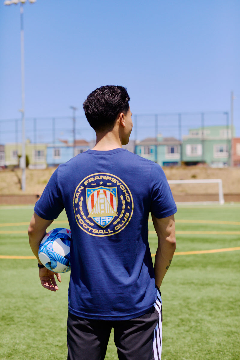 Man on a soccer field holding a ball, wearing the WC x SFP Tee in blue with a Team USA football club logo on the back.