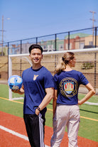 Two people are on a soccer field in San Francisco; one holds a ball and smiles, while the other faces away, proudly showing the WC x SFP Tee.