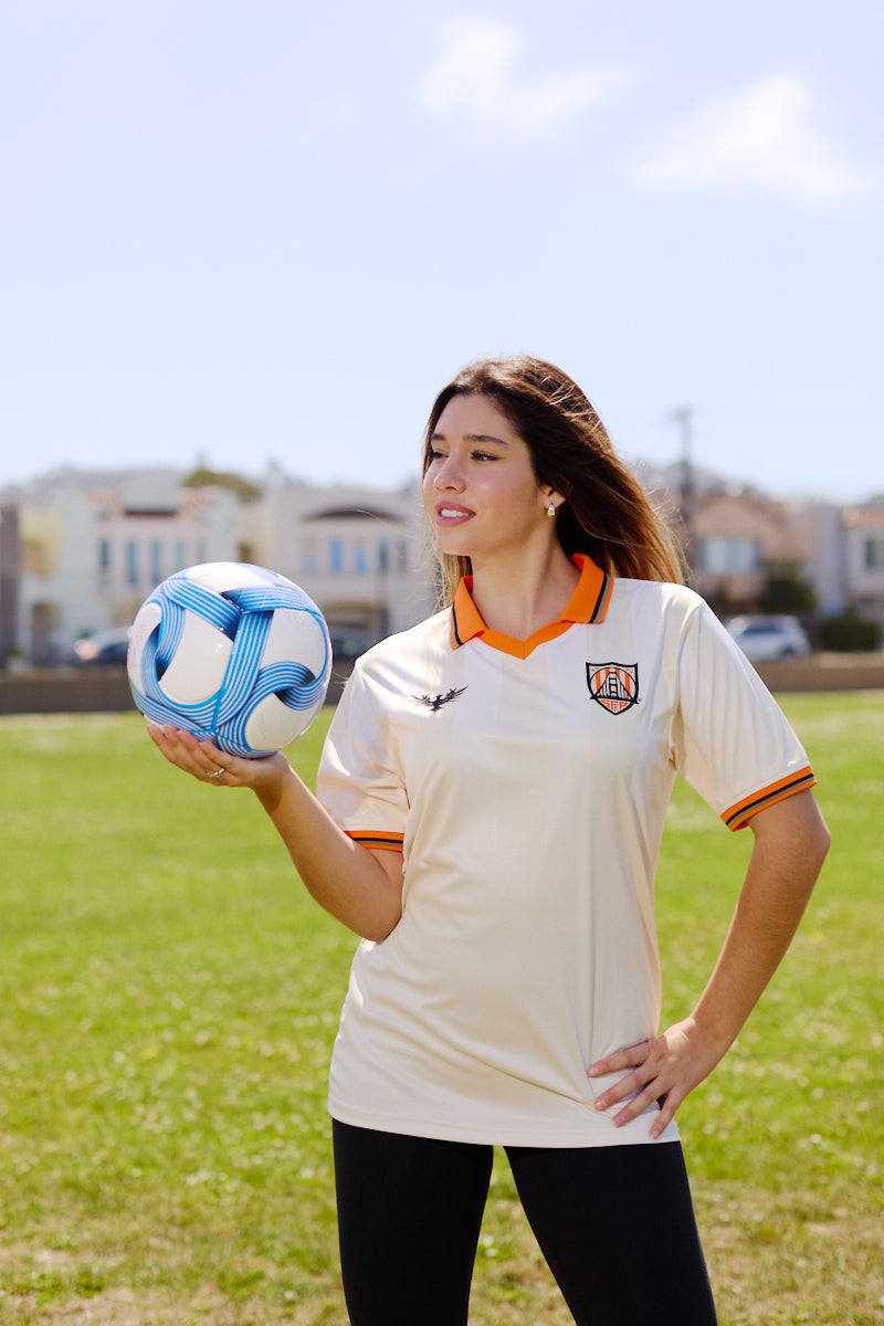 A woman wearing the WC x SFP Jersey - Away holds a blue and white soccer ball on a grassy field.