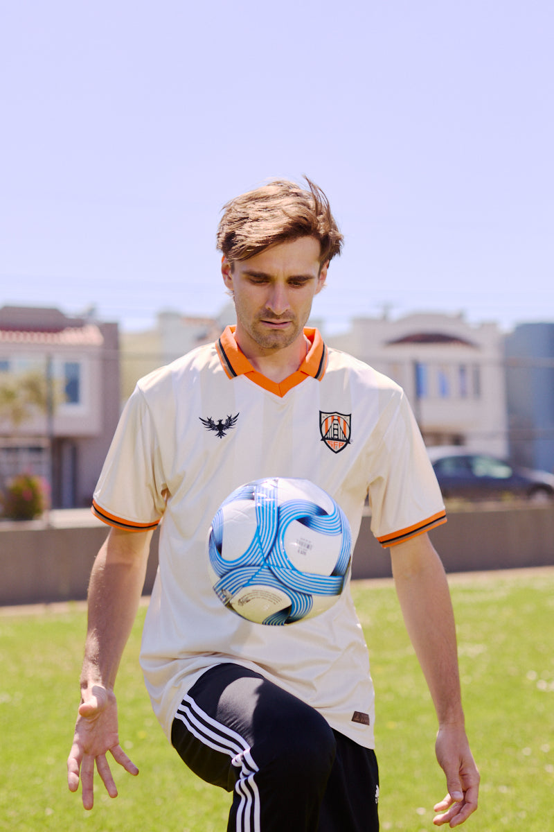 A man wearing the WC x SFP Jersey - Away juggles a blue and white soccer ball outdoors on a sunny day.