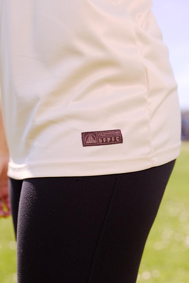 Person wearing the WC x SFP Jersey - Away, a cream shirt with a small patch and fan fit style, paired with black leggings, standing on grass outdoors.