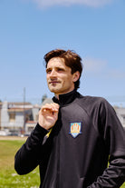 A man wears a WC x SFP Zip-Up Jacket with a sports logo while standing outdoors on a sunny day.