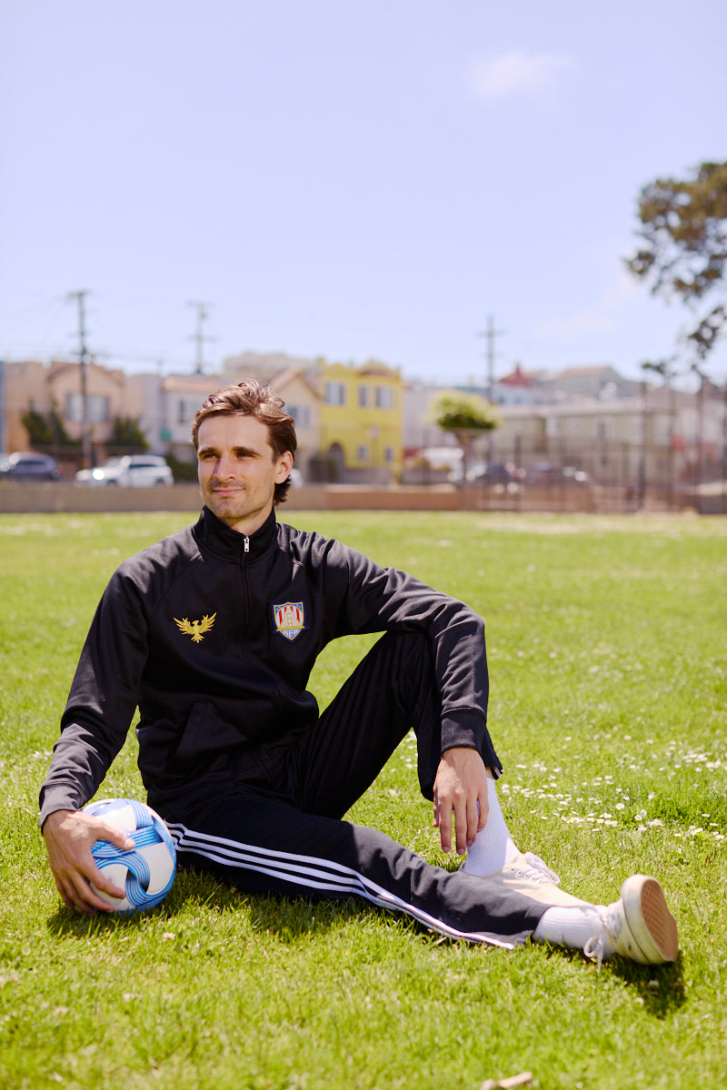 A man wearing the WC x SFP Zip-Up Jacket sits on grass with a soccer ball under the sun, capturing the US Men's National Team spirit in a park.